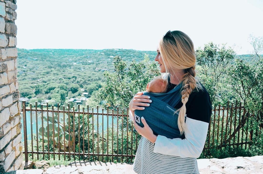 A blonde woman with a braided hairstyle holds a baby in a blue wrap carrier while looking out over a scenic, green landscape.