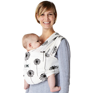 A person wearing a white Baby K'tan baby carrier with a dandelion pattern, carrying a sleeping infant.