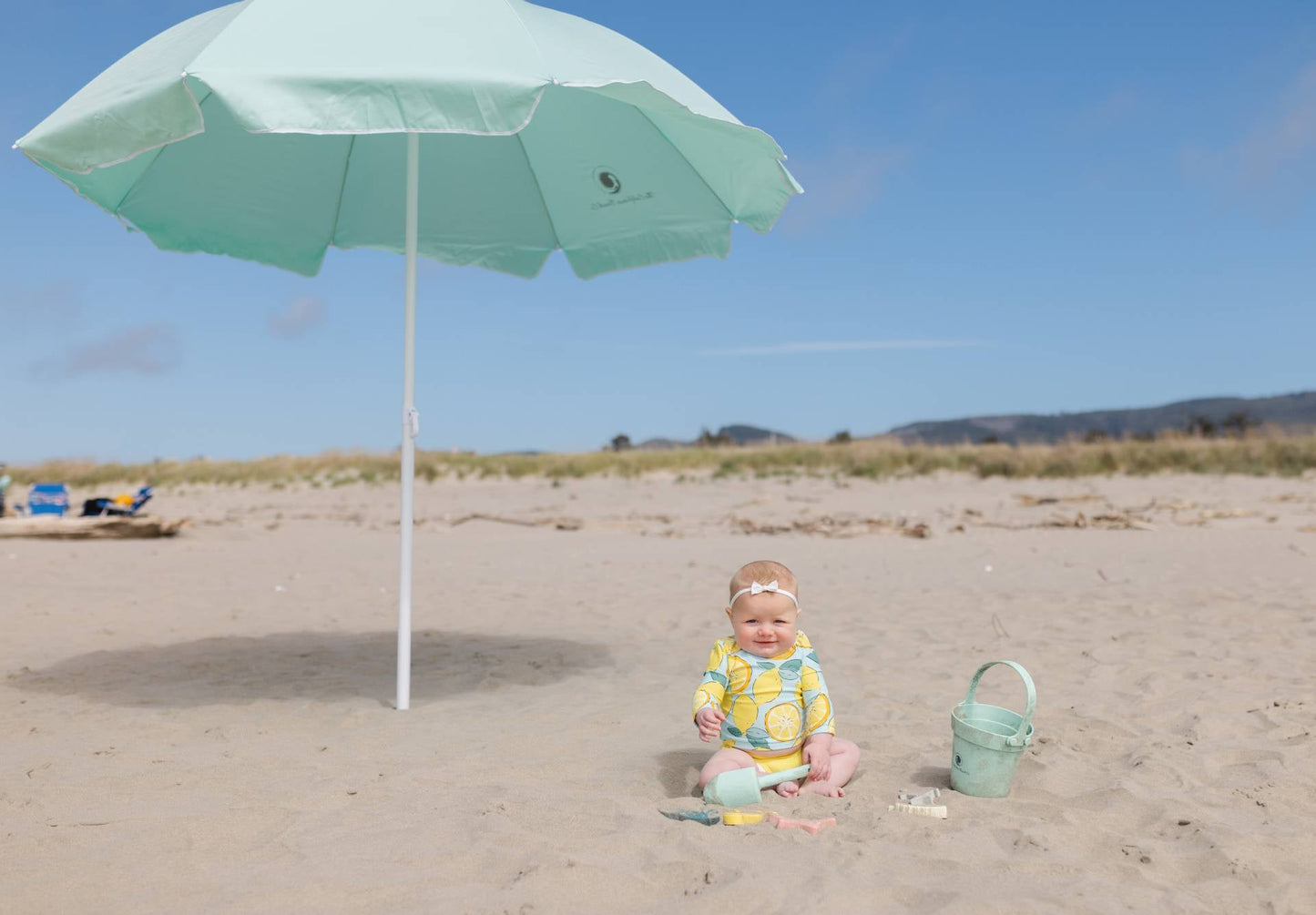 Baby on a sandy beach with a large turquoise umbrella and a bucket.