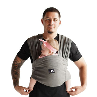 A man wearing a Baby K'tan carrier, holding a baby close to his chest with a gentle smile.