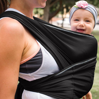 Woman in athletic wear carries a smiling baby in a black wrap. Baby wears a headband with a pink flower. Outdoor setting, joyful mood.