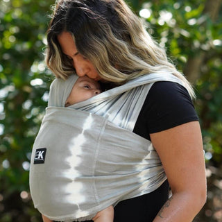 A woman with a baby in a grey Baby K'tan wrap carrier, showcasing a practical and stylish way to carry infants.