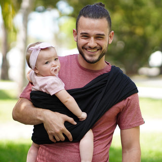 A man wearing a black baby carrier holds a baby close to his chest, showcasing a Baby K'tan design.