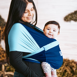 A woman smiles gently at a baby wrapped in a light and dark blue baby sling. The baby, wearing patterned leggings, looks towards the camera happily.