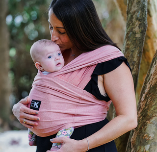 A woman affectionately kisses a baby wrapped in a pink carrier against her chest. They stand outdoors with trees in the background, conveying warmth and love.