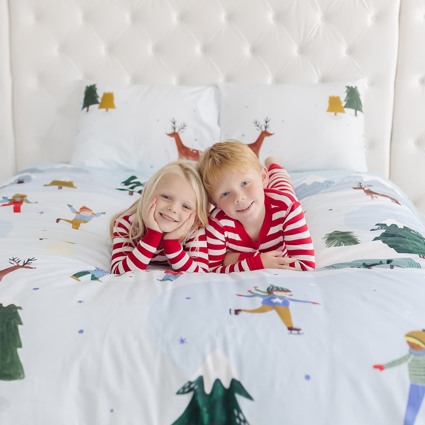Two children in matching red and white pajamas lying on a bed with a Christmas-themed duvet cover.