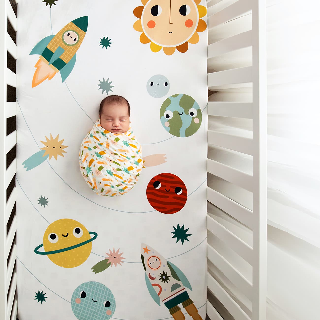 Baby swaddled in a colorful blanket with space-themed illustrations in a crib.