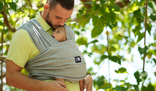 A man wearing a baby carrier walks through a wooded area, surrounded by trees and natural scenery.