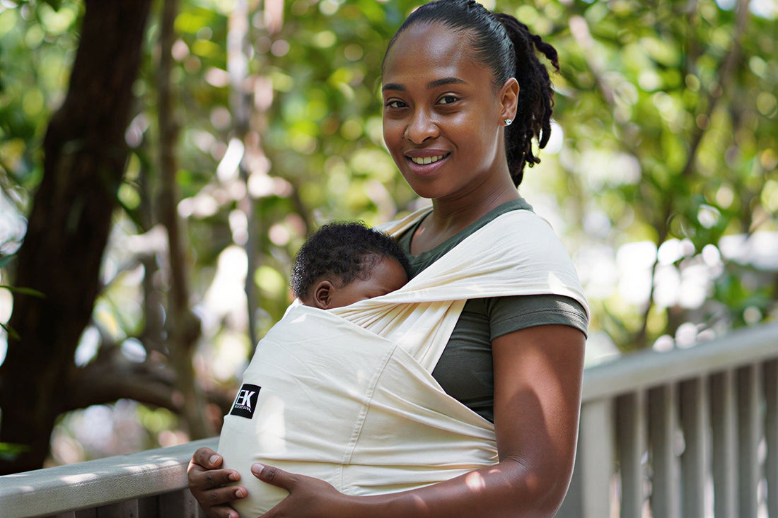 A smiling mother carrying her sleeping infant in a cream-colored fabric baby wrap while outdoors.