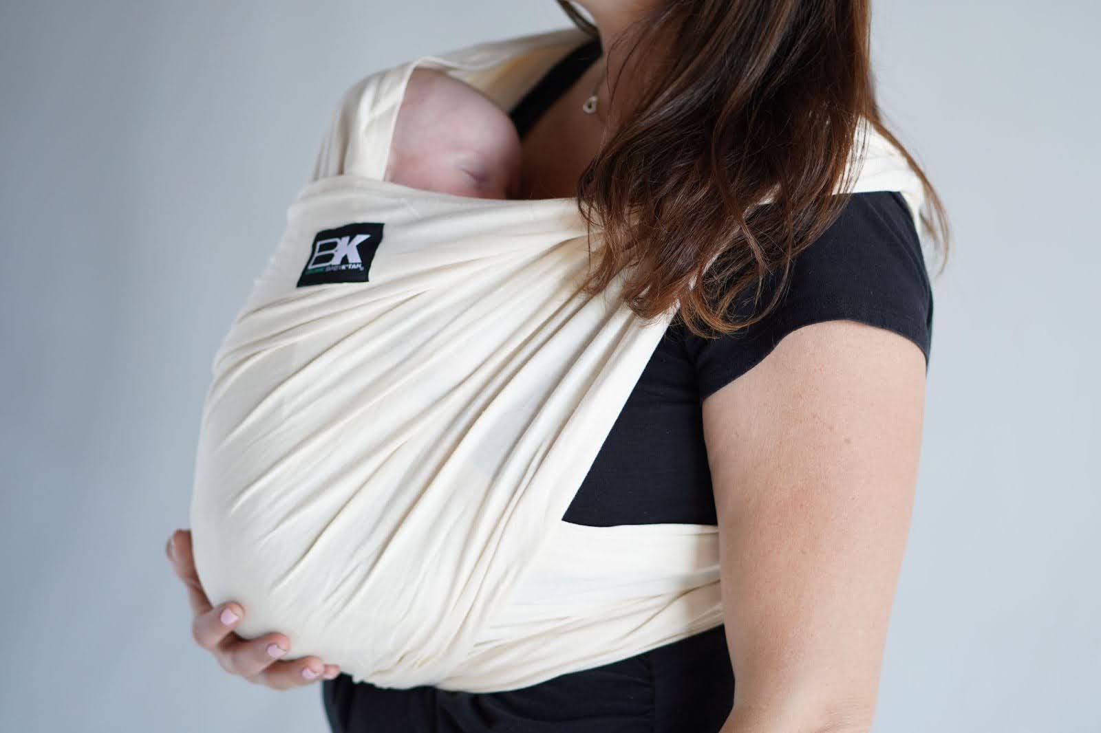 A woman in a black shirt carries a baby securely in a cream baby wrap against a neutral background