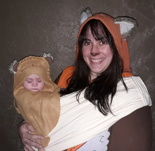 A woman in a colorful costume holds a baby in a carrier, smiling warmly at the camera.
