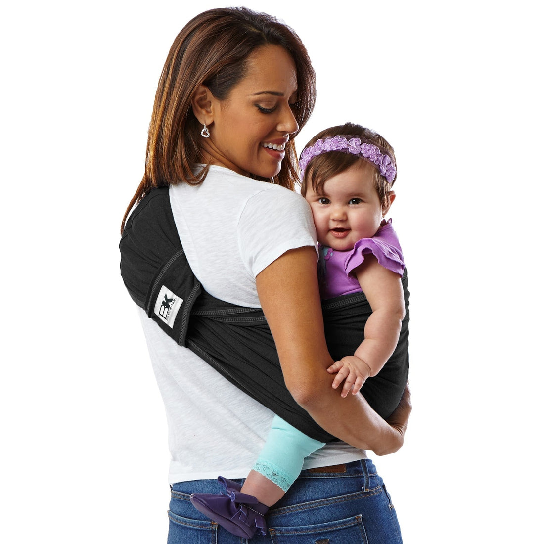 A woman wearing a white shirt and jeans carries a baby in a black baby sling, with the baby dressed in a purple outfit and headband.