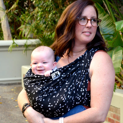 A woman with glasses lovingly holds a smiling baby in a black patterned sling. They are outside, surrounded by lush green plants, creating a warm, caring atmosphere.