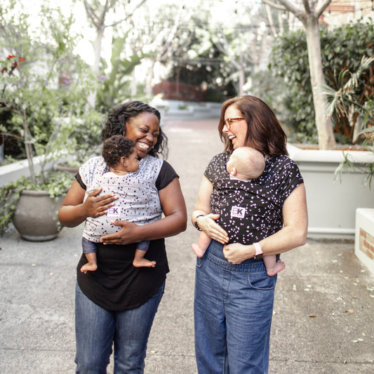 A woman is wearing a Baby K'tan Baby Carrier in Sweetheart Grey pattern with a smiling baby inside it. She is holding the carrier up to the camera, and they both appear happy.