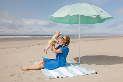 Woman and child under a green beach umbrella on a sandy beach.