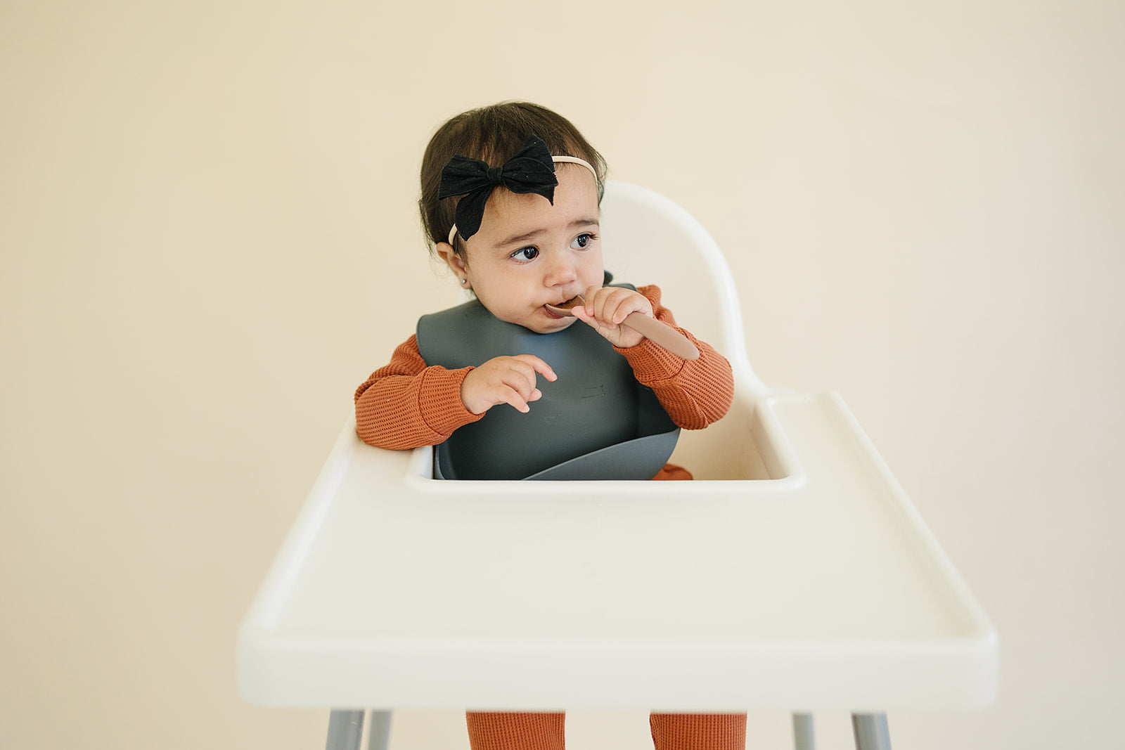 Child in a high chair with a bib, eating with a spoon against a beige background