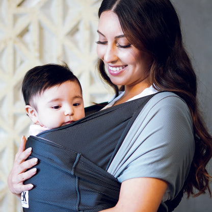 A smiling woman holds a baby in a black carrier against a patterned wall. The scene conveys warmth and contentment, with the baby looking calm and cozy.