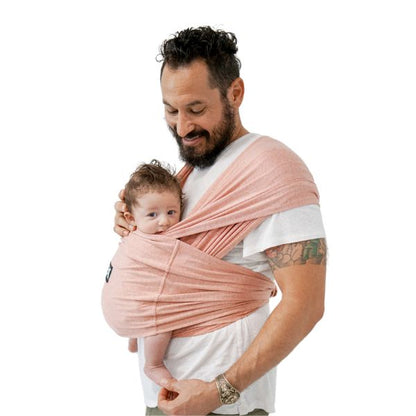 Smiling man holding a baby in a pink wrap. The infant looks content and curious. The man, with a tattooed arm, exudes warmth and care.