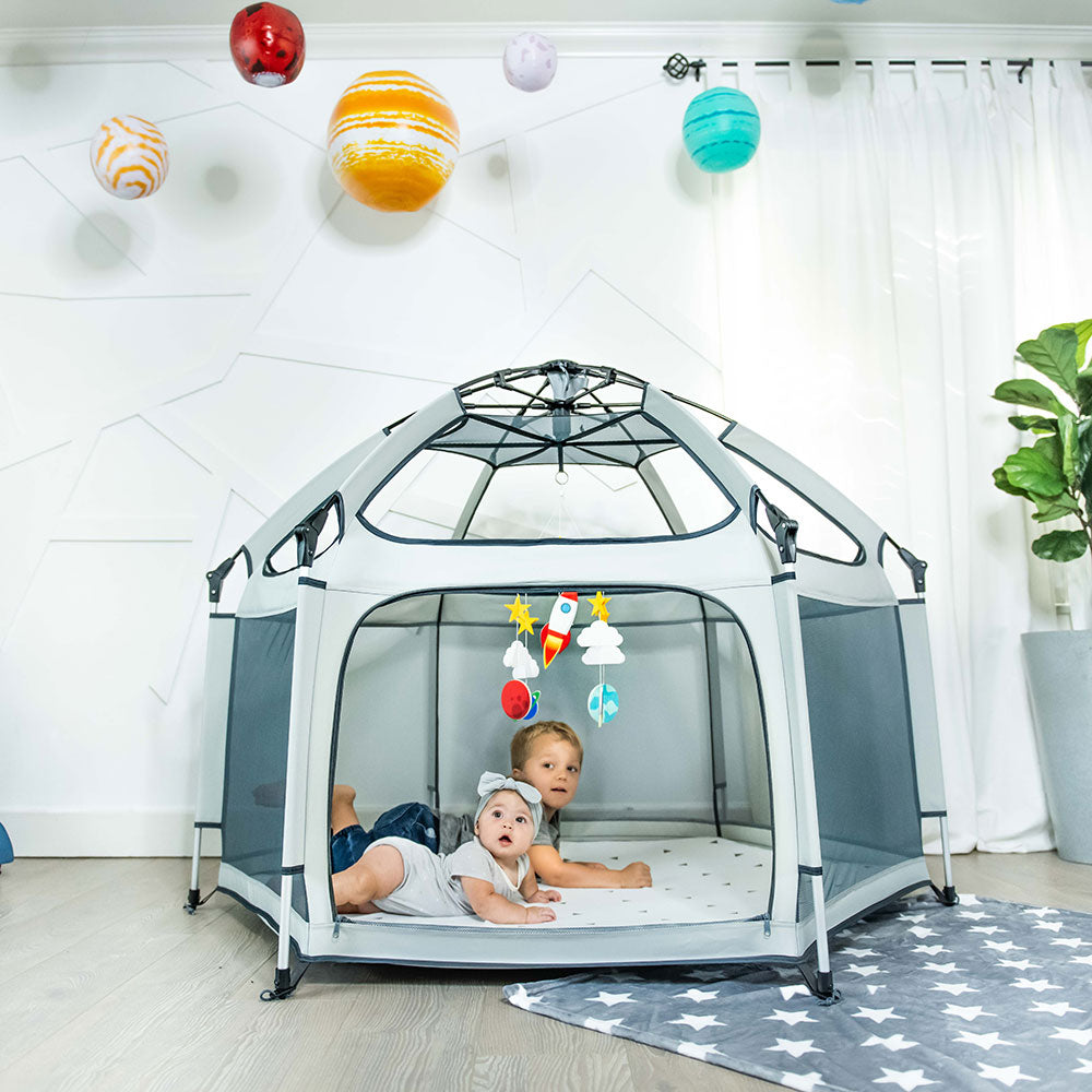 Children playing inside a dome-shaped playpen with colorful toys hanging above.