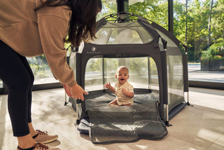 Baby sitting inside a portable playpen with a person adjusting it, in an indoor setting.