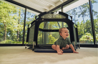 Baby crawling on a wooden floor next to a portable playpen with trees outside.