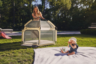 Woman standing inside a portable tent on grass with a child sitting outside.