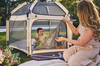 Woman setting up a portable playpen with a child inside in a grassy outdoor area.