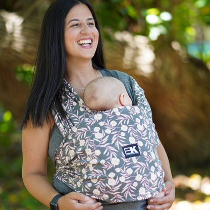 A woman with a baby K'tan carrier is seen walking in a serene forest, surrounded by greenery and dappled sunlight.