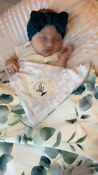 Newborn baby wrapped in a blanket with a visible brand logo, wearing a dark blue headband, on a textured surface.