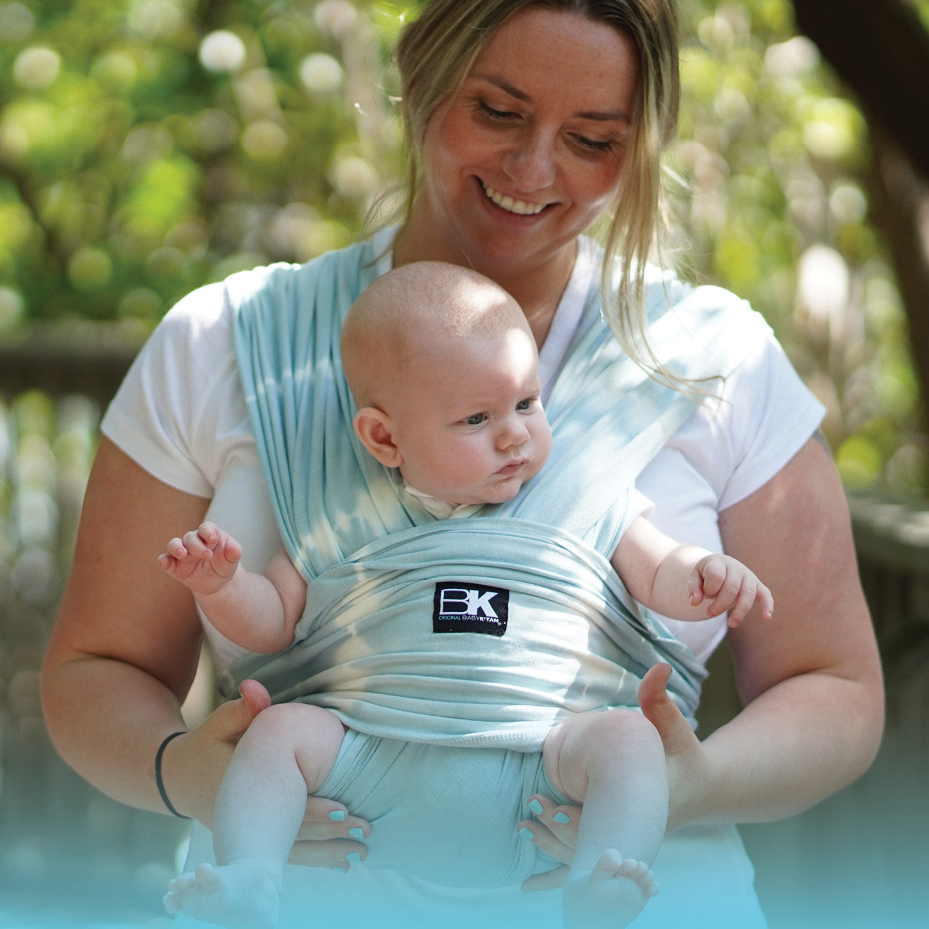 A smiling woman in a white shirt holds a baby wrapped in a light blue baby carrier outdoors. The scene conveys warmth and contentment.