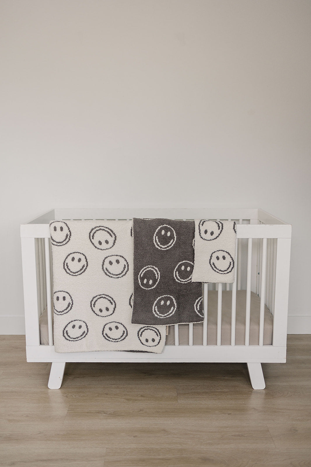 White crib with smiley face patterned blanket on a wooden floor.
