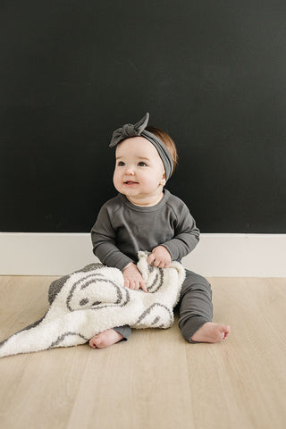 Baby sitting on a wooden floor with a black background, holding a blanket.