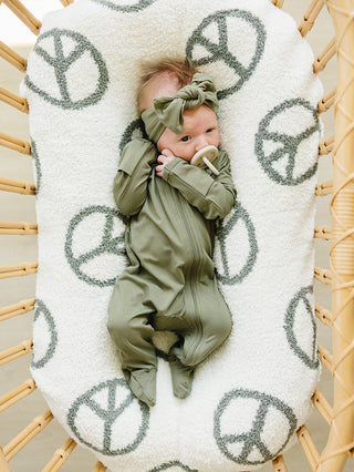 Baby in a green outfit lying on a white blanket with peace symbol patterns in a wicker crib.
