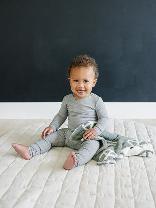 Baby in a gray outfit sitting on a bed with a dark blue wall in the background