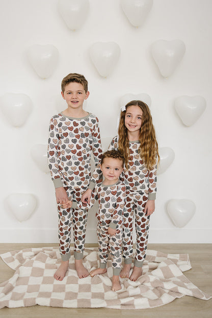 Three children wearing matching pajamas with a heart pattern, standing on a checkered blanket against a white wall with heart shapes.