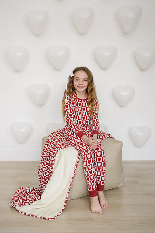 Young girl wearing a red and white patterned onesie sitting on a beige ottoman with a heart-shaped balloon background.