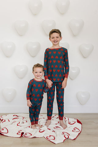Two children wearing red heart-patterned pajamas standing on a white rug with heart designs against a white wall.