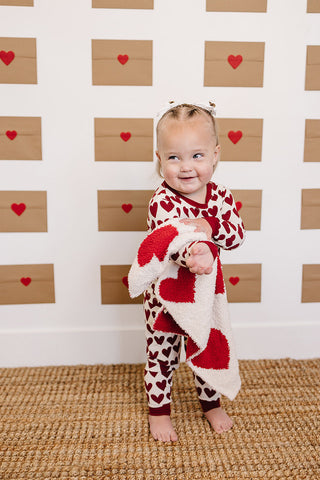 Child wearing a heart-patterned outfit with a red and white heart-shaped blanket against a wooden heart backdrop.