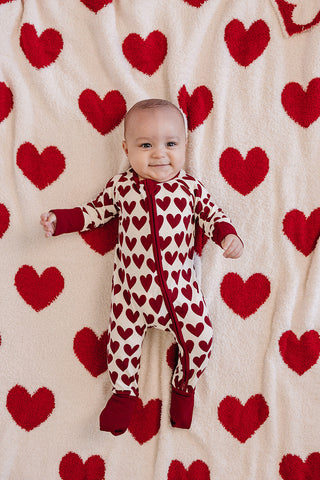 Baby in a heart-patterned onesie standing against a backdrop of red hearts on a white background