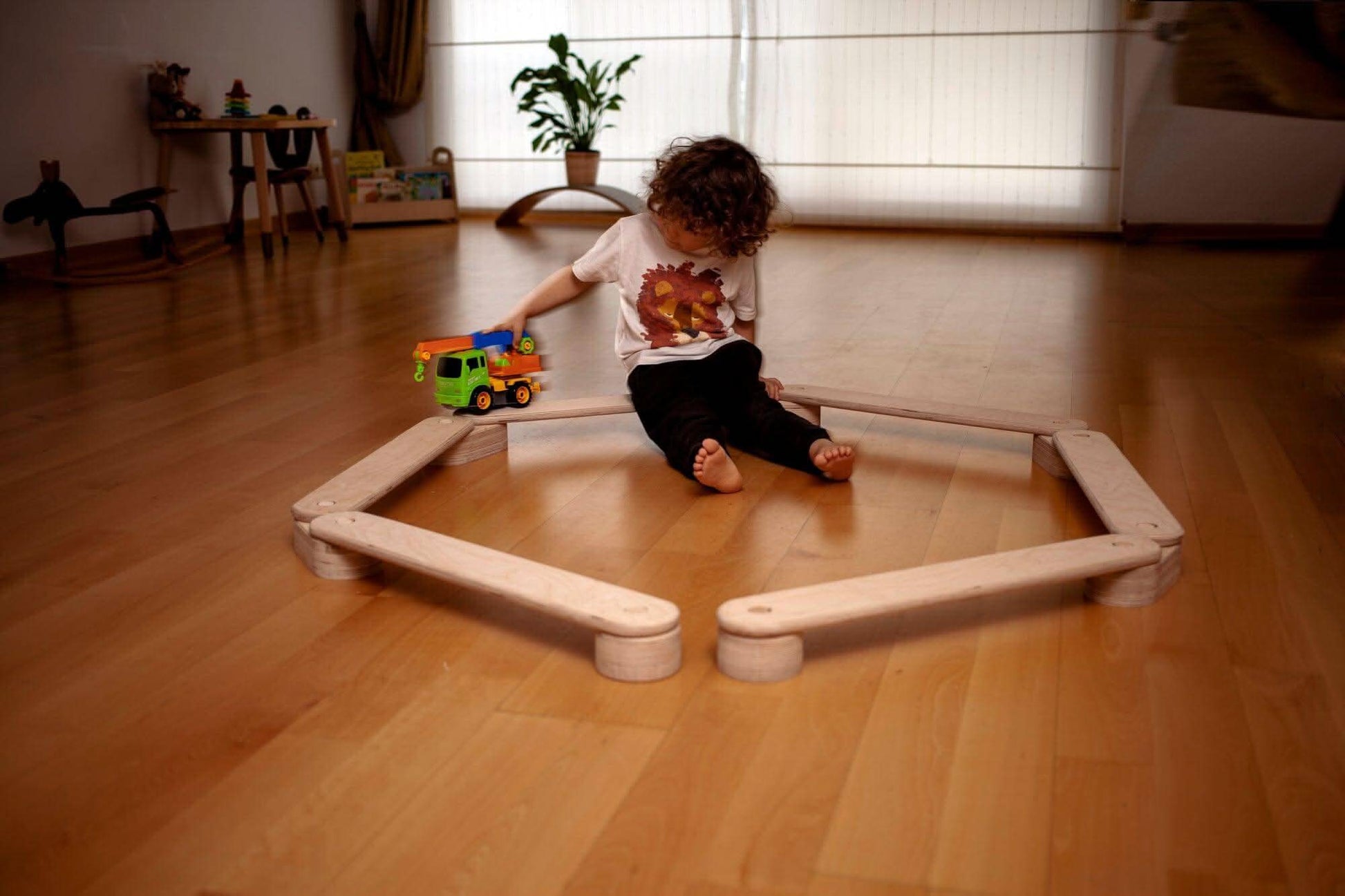 Child playing with a toy truck on a wooden floor in a room with a window and plant.