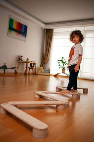 Child playing with wooden balance beams in a room with a desk and plants.