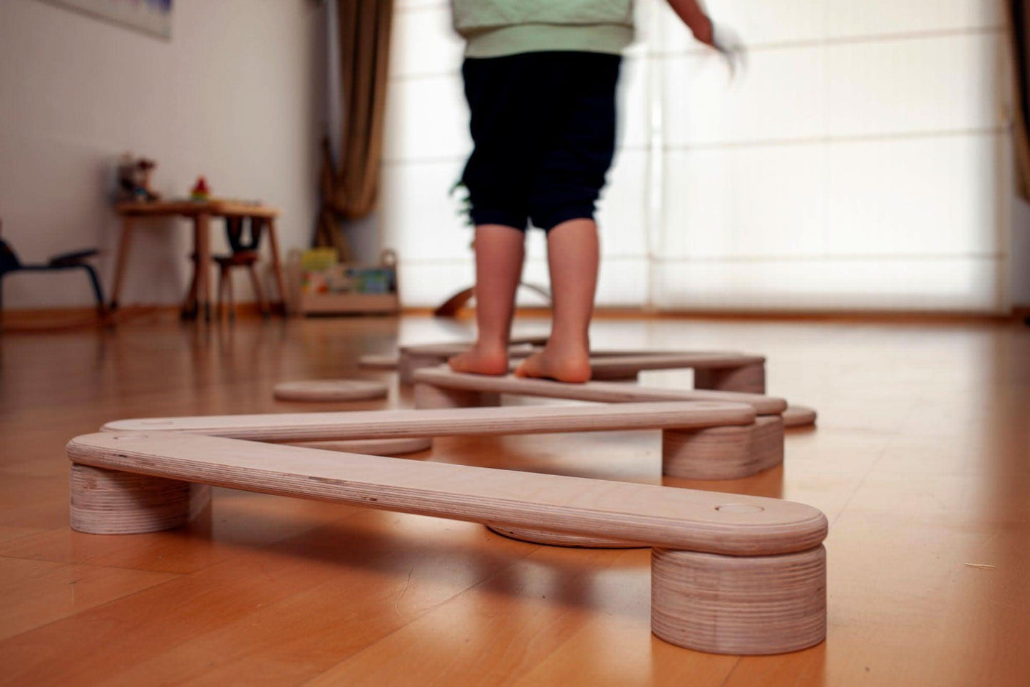 Child standing on a wooden balance beam in a room with wooden flooring.