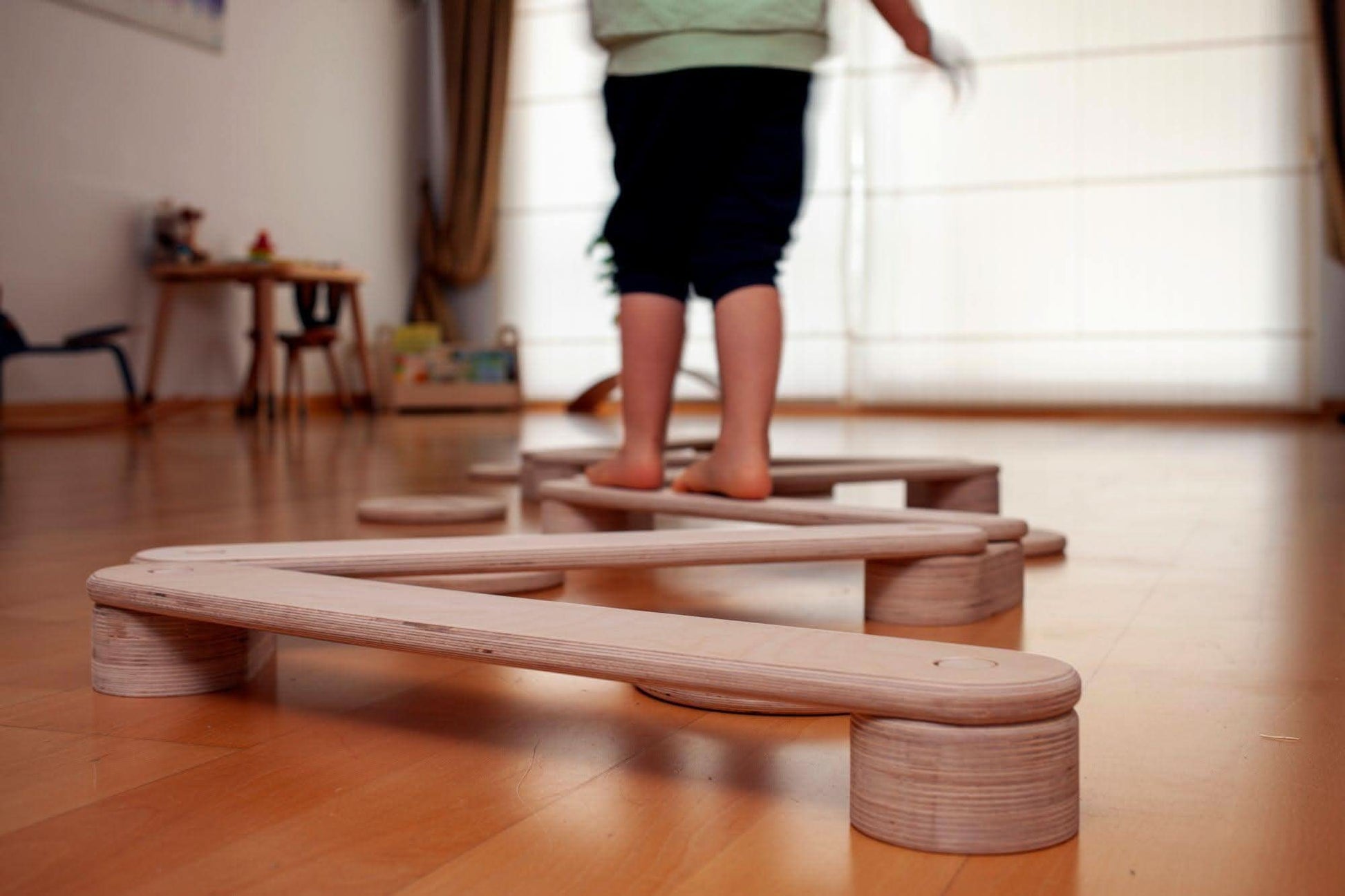 Child standing on a wooden balance beam in a room with wooden flooring.