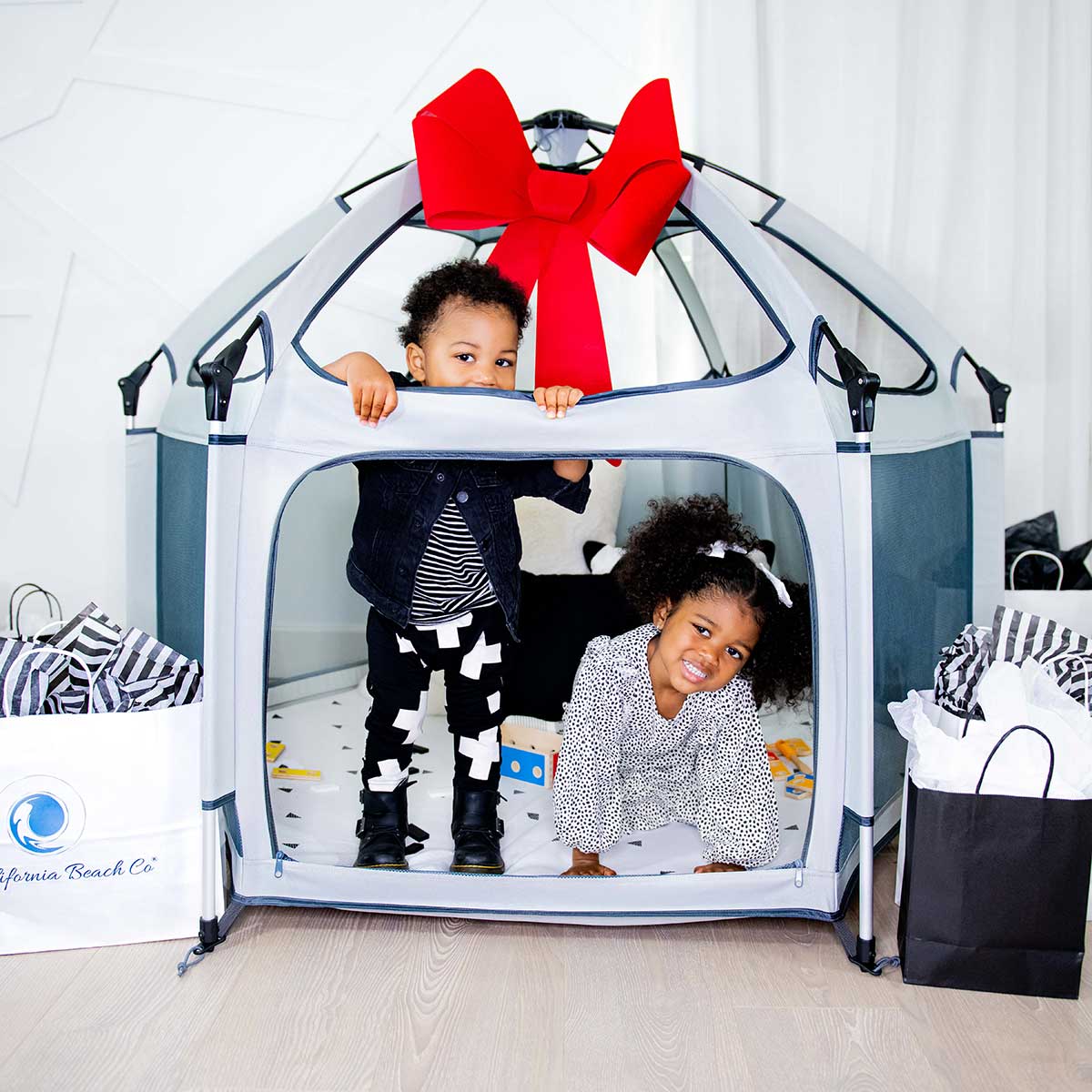 Two children playing inside a play tent with a large red bow, surrounded by shopping bags.