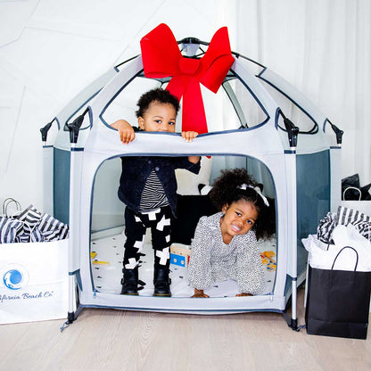 Two children playing inside a play tent with a large red bow, surrounded by shopping bags.