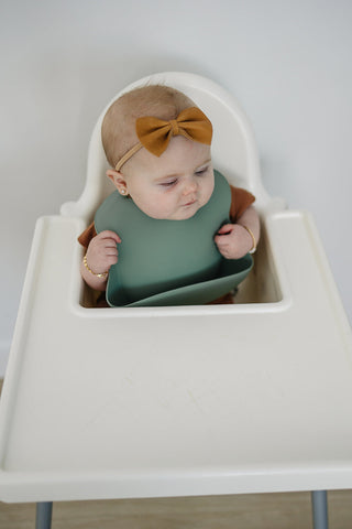 Baby in a high chair wearing a green bib and brown bow