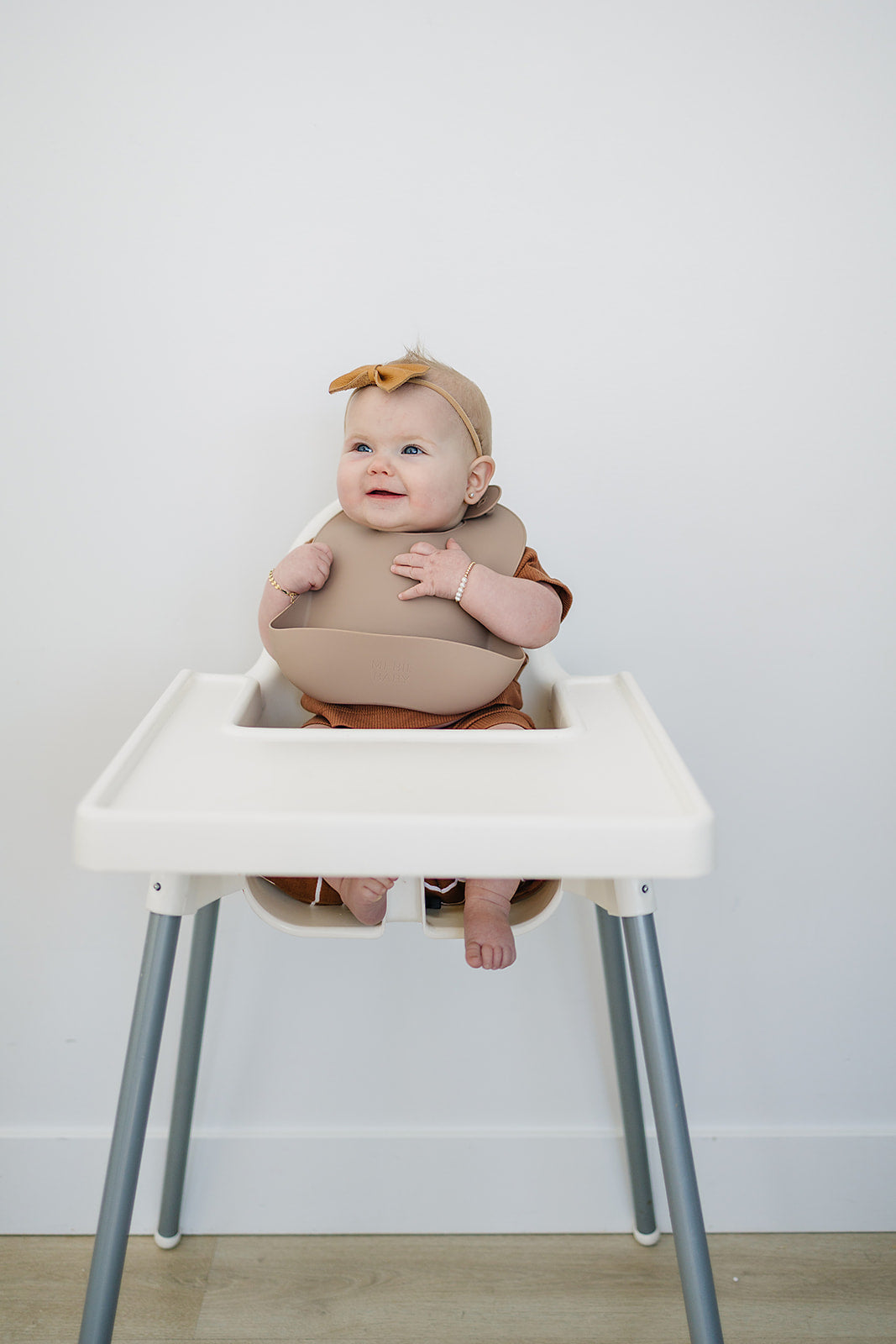 Baby sitting in a high chair wearing a brown outfit and headband against a white wall.
