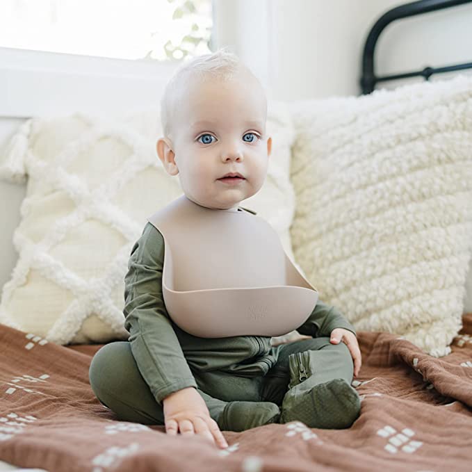 Baby wearing a green outfit with a beige bib sitting on a bed.