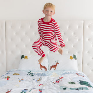 Child in red and white striped pajamas jumping on a bed with a festive comforter.