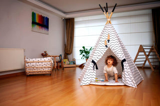 Child playing inside a teepee in a room with wooden floor and white walls.